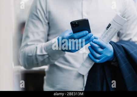 Cropped shot of faceless man dressed in white shirt, medical rubber gloves to prevent coronavirus, uses mobile phone, downloads new app, holds newspap Stock Photo