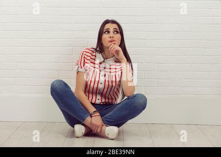 Thinking young woman sitting cross-legged, looking up, isolated white brick wall background. Striped shirt, jeans. Neutral human emotion facial expres Stock Photo