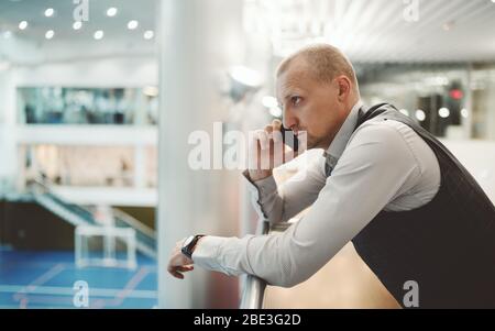 A serious robust hispanic man entrepreneur is having a phone conversation while leaning against chrome railing of a balcony in a contemporary office i Stock Photo