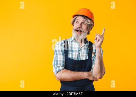 Photo of elderly grey-haired bearded man builder in helmet posing isolated over yellow wall background have an idea. Stock Photo