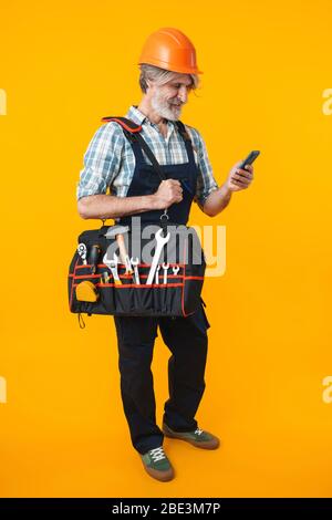 Image of cheery elderly grey-haired bearded man builder in helmet posing isolated over yellow wall background holding bag with equipment instruments u Stock Photo