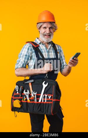 Image of optimistic happy elderly grey-haired bearded man builder in helmet posing isolated over yellow wall background holding bag with equipment ins Stock Photo