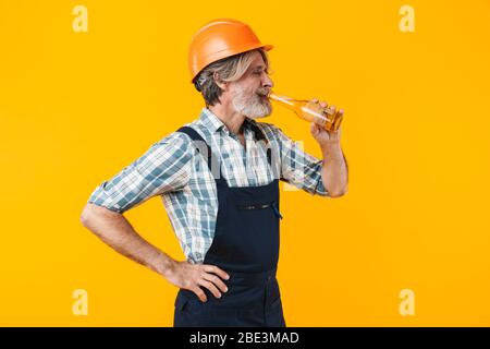 Image of positive elderly grey-haired bearded man builder in helmet posing isolated over yellow wall background drinking beer. Stock Photo