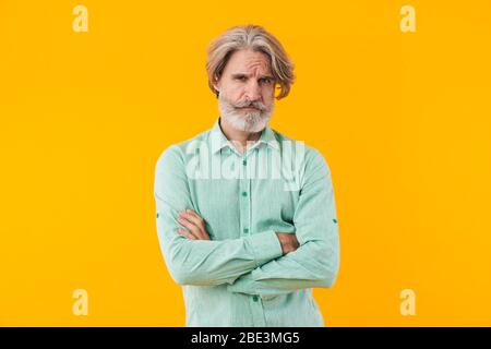 Image of serious negative elderly grey-haired bearded man in blue shirt posing isolated over yellow wall background. Stock Photo