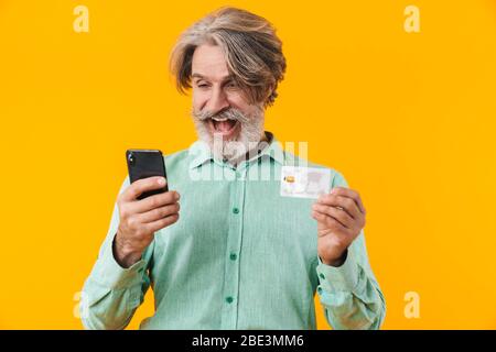 Photo of surprised grey-haired bearded man in blue shirt posing isolated over yellow wall background using mobile phone holding credit card. Stock Photo