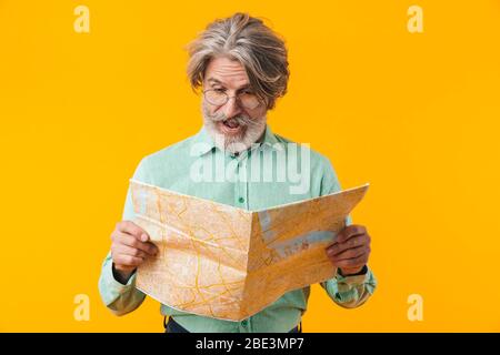 Picture of shocked grey-haired man posing isolated over yellow wall background holding map. Stock Photo