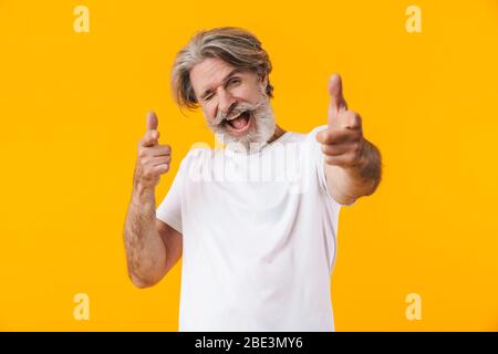 Image of emotional cheerful senior grey-haired bearded man posing isolated over yellow wall background pointing to you. Stock Photo