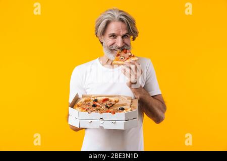 Image of positive smiling senior grey-haired bearded man posing isolated over yellow wall background eat pizza. Stock Photo