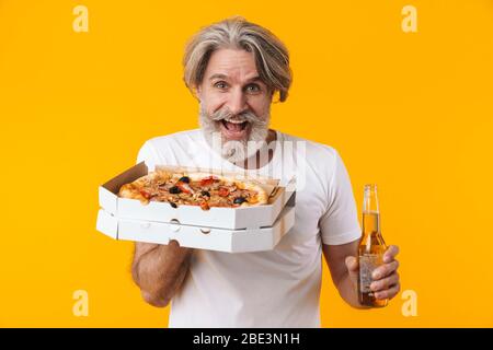 Photo of surprised emotional senior grey-haired bearded man posing isolated over yellow wall background holding pizza drinking beer. Stock Photo