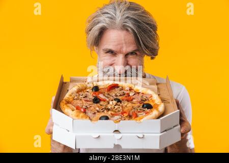 Image of positive smiling senior grey-haired bearded man posing isolated over yellow wall background eat pizza. Stock Photo