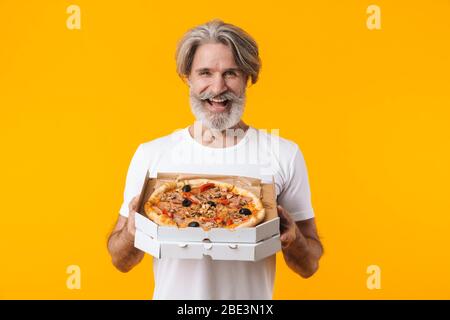 Image of positive smiling senior grey-haired bearded man posing isolated over yellow wall background eat pizza. Stock Photo
