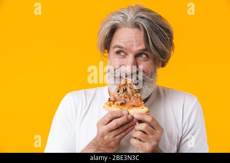 Image of happy funny emotional senior grey-haired bearded man posing isolated over yellow wall background holding pizza. Stock Photo