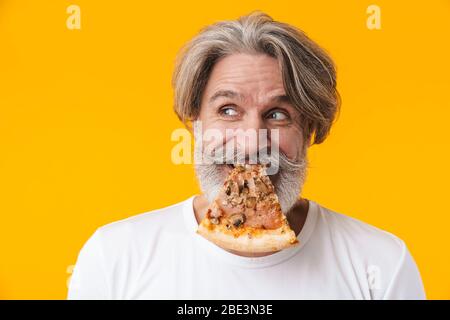Photo of positive funny senior grey-haired bearded man posing isolated over yellow wall background eat pizza. Stock Photo