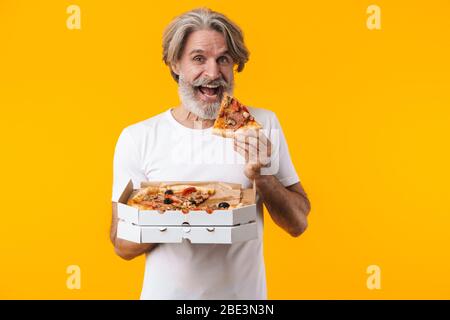 Image of positive smiling senior grey-haired bearded man posing isolated over yellow wall background eat pizza. Stock Photo