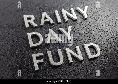 the words rainy day fund laid with silver metal letters on black background, all covered with water drops, slanted diagonal perspective with selective Stock Photo