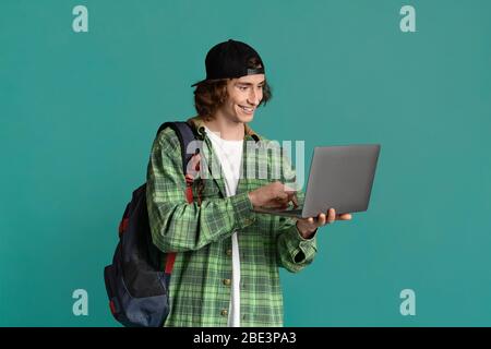 Online education. Teenage student with backpack using laptop for e-learning, turquoise background Stock Photo