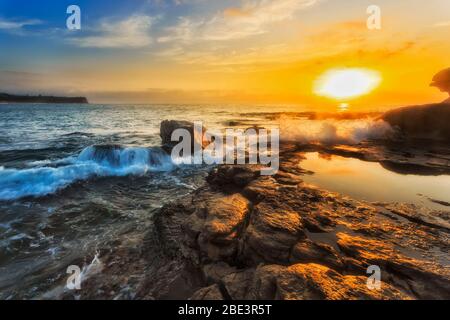 Sun over pacific ocean horizon off Turimetta head on Sydney Northern beaches with wave rolling on sandstone rocks.