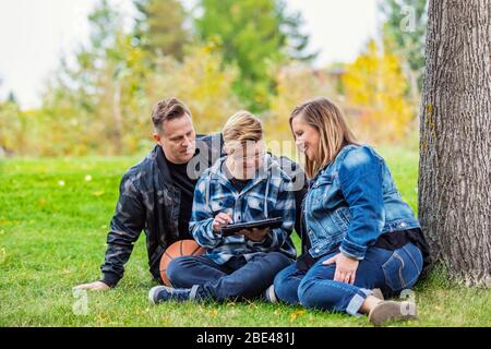 A young man with Down Syndrome learns a new program on a tablet with his father and mother while enjoying each other's company in a city park on a ... Stock Photo