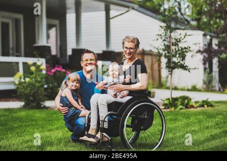 A young family posing for a family portrait outdoors in their front yard and the mother is a paraplegic in a wheelchair; Edmonton, Alberta, Canada Stock Photo
