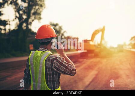 Engineer wear a hard hellmet and usingsmartphone on road construction site with machinery,safety first concept Stock Photo