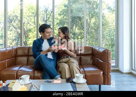Young couples spend holidays in the living room. The young man wears comfortable clothes, hand a gift box  to surprise girlfriend on special occasions Stock Photo