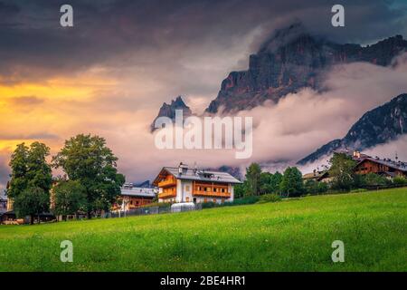 Charming alpine village with flowery green fields and high misty mountains in background at sunset, Dolomites, San Vito di Cadore, Italy, Europe Stock Photo