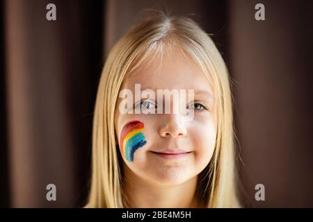 Girl with painted rainbow on face during Covid-19 quarantine at home. Stay at home Social media campaign for coronavirus prevention, let's all be well Stock Photo