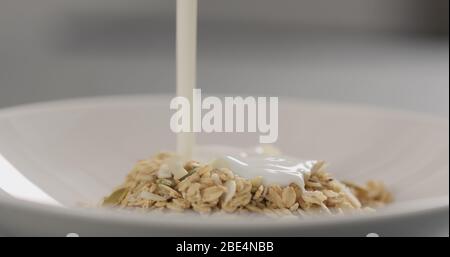 pour yogurt over granola with almond flakes and pumpkin seeds into white bowl closeup Stock Photo