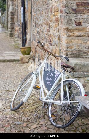 Old fashioned white painted women's bicycle propped up against a pale yellow stone exterior wall outside a café in a pretty village location Stock Photo