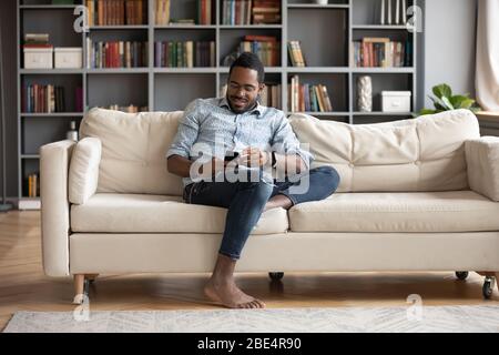 Smiling young african man sitting on comfy couch, playing game. Stock Photo