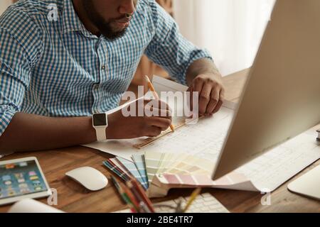 Architect or designer working at home. Cropped photo of young afro american man drawing something on blueprint while sitting at his working place at home. Freelance. Home office workplace Stock Photo