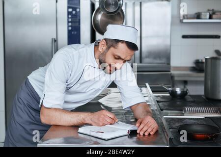 Concentrated male chef cook with grocery list or bills doing inventory at restaurant kitchen. Cooking and people concept. Side view Stock Photo