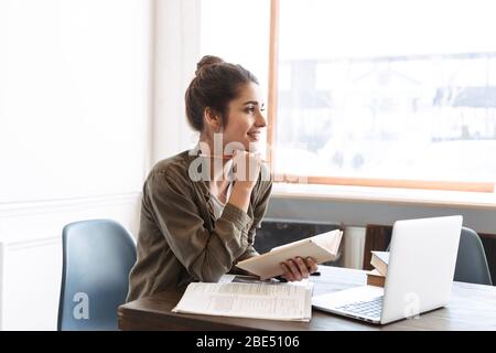 Image of a beautiful happy young concentrated woman using laptop computer indoors writing notes in notebook reading book. Stock Photo