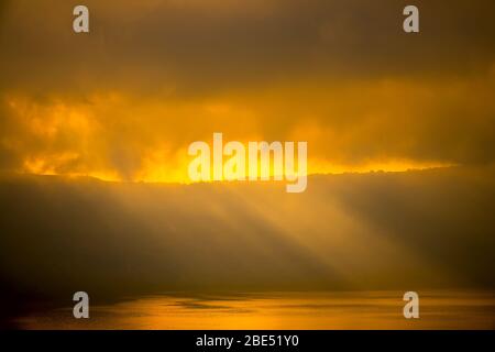 Heavy clouds over the rocky shore. Sunrays barely break through, illuminate houses and the water surface Stock Photo
