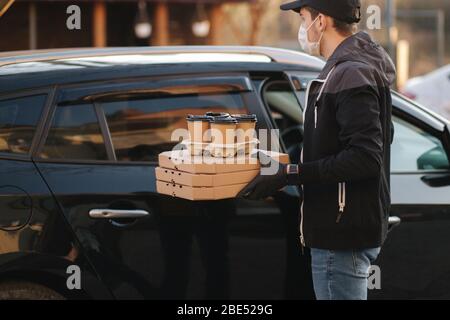 Courier in protective mask and medical gloves take order from car. Delivery boy hold pizza and coffe. Delivery service under quarantine. Coronavirus Stock Photo