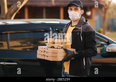 Courier in protective mask and medical gloves take order from car. Delivery boy hold pizza and coffe. Delivery service under quarantine. Coronavirus Stock Photo