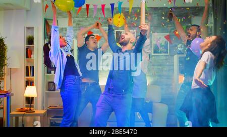 Attractive young man throwing confetti while partying with his group of friends in a room with neon lights, disco ball and alcohol. Slow motion shot Stock Photo