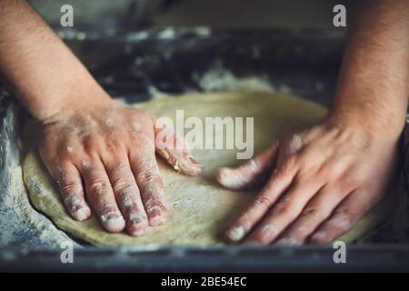 Man's strong hands roll out homemade pizza dough by hand, which lies on a dark baking sheet. Baking at home. Stock Photo