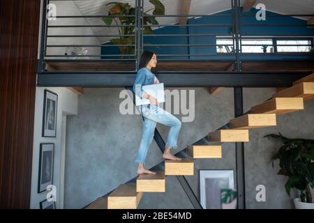 Young woman going upstairs, holding laptop under arm Stock Photo