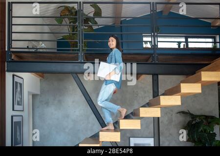Young woman going downstairs, holding laptop under arm, smiling Stock Photo