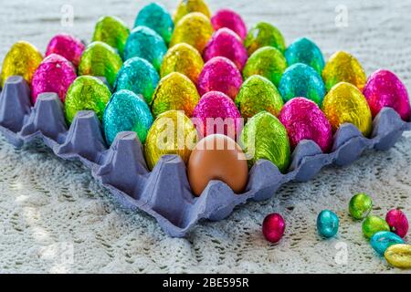 A hen egg among colorful Easter chocolate eggs in a carton. Stock Photo