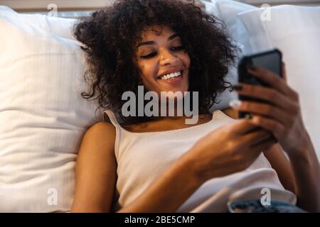 Photo of cheery young african woman indoors at home using mobile phone lies in bed. Stock Photo