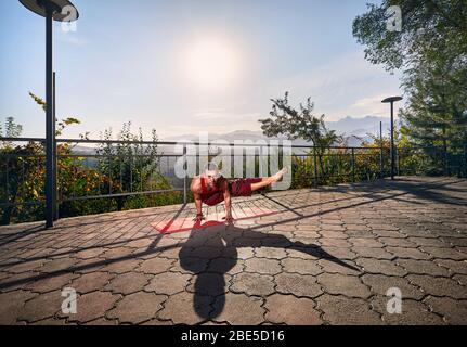 Fit man in red costume doing yoga bakasana pose in the park with mountain view in Almaty, Kazakhstan Stock Photo