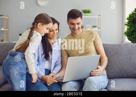 Happy family chatting online using laptop while sitting with baby on sofa at home. Stock Photo