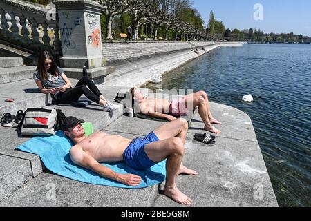 Konstanz, Germany. 12th Apr, 2020. Three people from Constance sunbathe on the promenade along the shore of Lake Constance, where there are hardly any boats on the water despite the beautiful weather. Many people use the sunny weather for an Easter walk. Credit: Felix Kästle/dpa/Alamy Live News Stock Photo