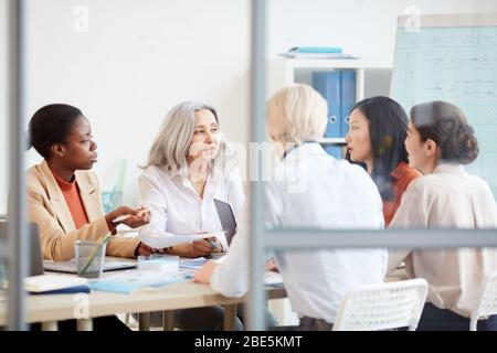 Portrait of diverse female business team discussing project while sitting at table during meeting in conference room, shot from behind glass wall Stock Photo