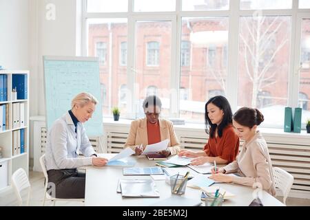 Diverse group of successful businesswomen discussing project while sitting at table against window during meeting in conference room, copy space Stock Photo