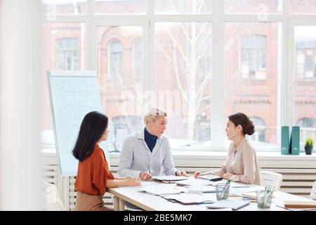 Group of modern businesswomen discussing project while sitting at table against window during meeting in conference room, copy space Stock Photo