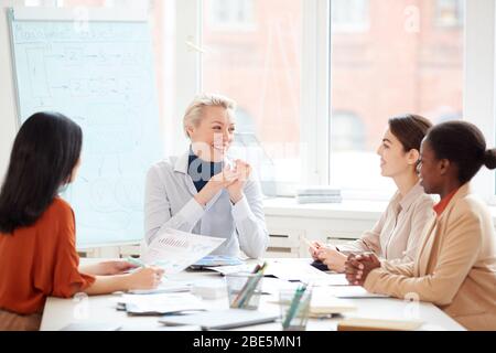 Group of cheerful businesswomen sharing ideas while sitting at table against window during meeting in conference room, copy space Stock Photo