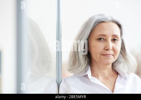 Head and shoulders portrait of long-haired senior businesswoman looking at camera while leaning against glass wall in office, left side copy space